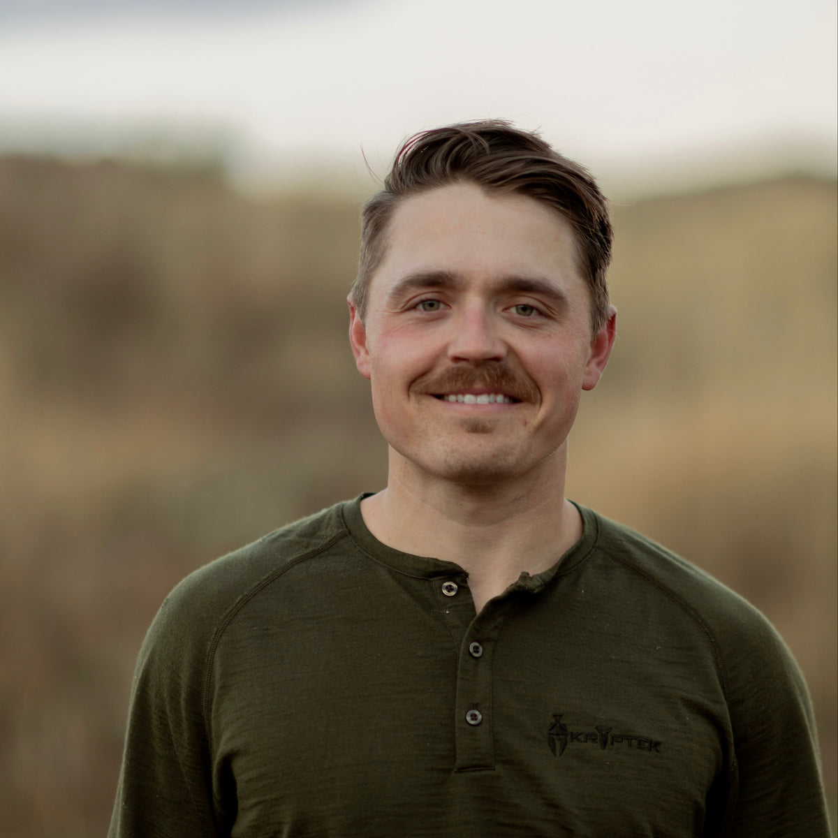 Man wearing a green long-sleeve shirt with a logo, standing outdoors with a blurred natural background